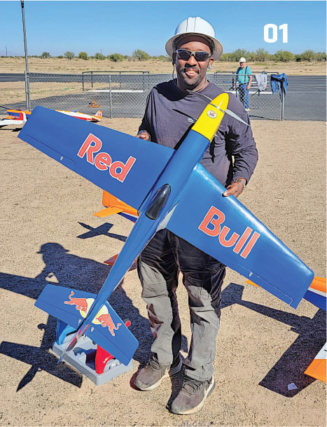 Man holding a blue model airplane with "Red Bull" text, standing on a sandy lot.