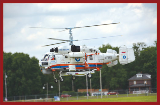 White and blue helicopter flying over a grassy field with trees in the background.