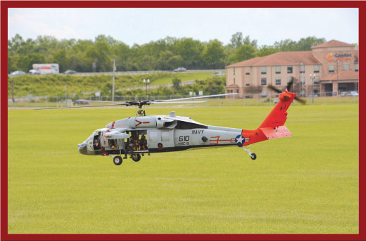 Navy helicopter with red tail flying low over grassy field.