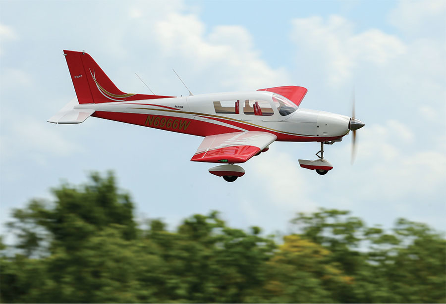 Red and white small airplane flying over blurred trees, clear sky background.