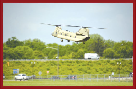 Tan military helicopter flying above a grassy field, with trees and vehicles in the background.
