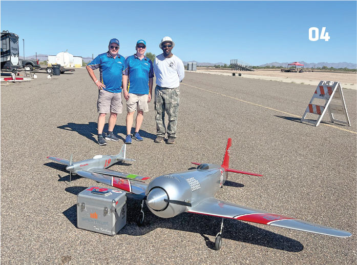 Three men stand on airstrip with a large model plane and a silver box.