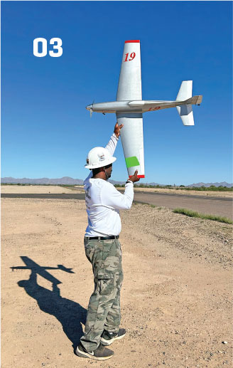 Man in camo pants holding a model airplane, number 19, under a clear blue sky.