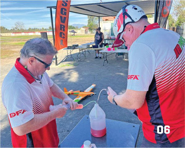 Two men in red and white shirts preparing model aircraft equipment outdoors.