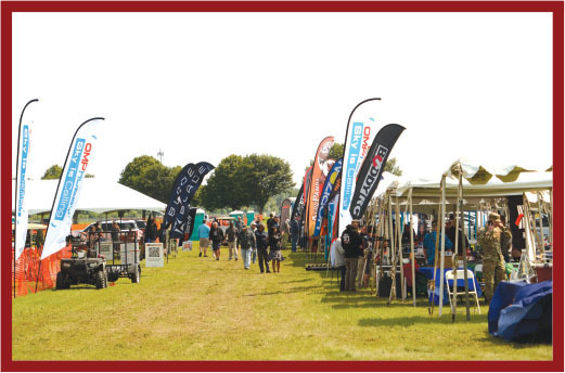 Outdoor event with tents, people, and colorful flags on a grassy field.