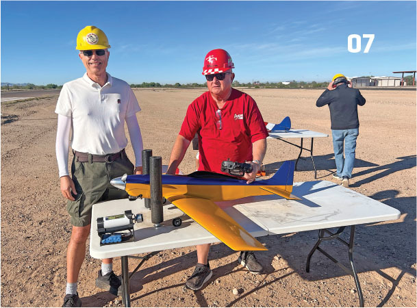 Two men in hard hats with a model airplane on a table outdoors.