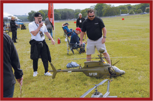 Model helicopters on grass, two men observing, one smiling.
