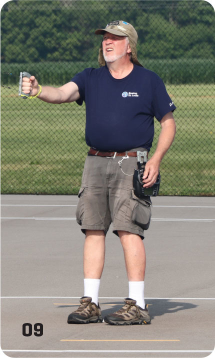 Man in shorts and hat on a court, holding a stopwatch with a green field behind.