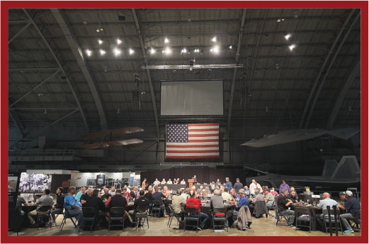 Large gathering in a hangar with U.S. flag displayed, people seated at round tables.