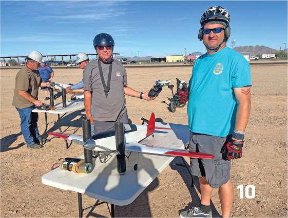 Men wearing helmets beside model airplanes on outdoor tables.
