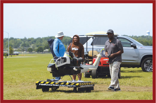 Three men outdoors with a hovercraft and vehicles in a grassy area.