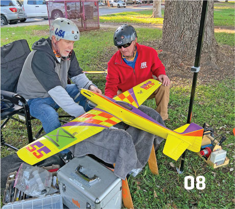 Two men with helmets adjust a colorful model plane outdoors.