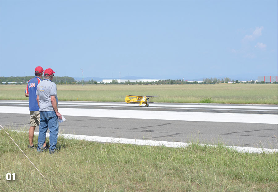 Two men in red caps watch yellow model plane on grass runway.