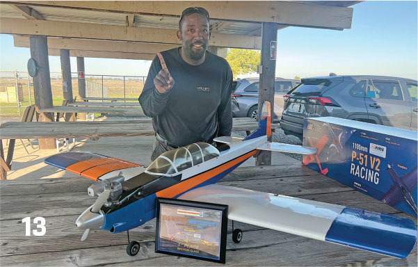Man smiling, holding up one finger, beside a model airplane on a wooden bench.