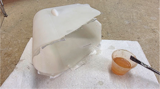 Broken white plastic tank on a mat, next to a cup of resin and a brush.