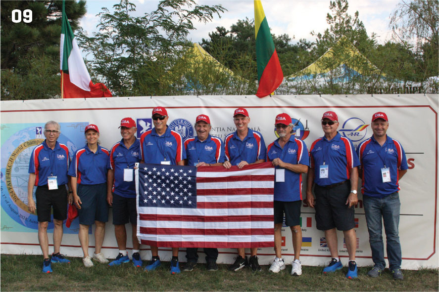 Team wearing blue shirts and red hats holding a U.S. flag in front of banners and flags.