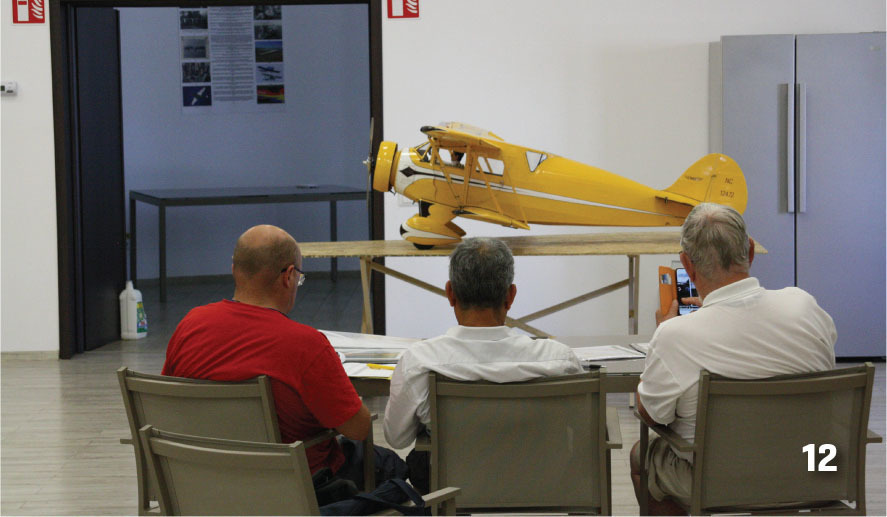 Three men seated, observing a yellow model airplane on a table, number "12" in corner.