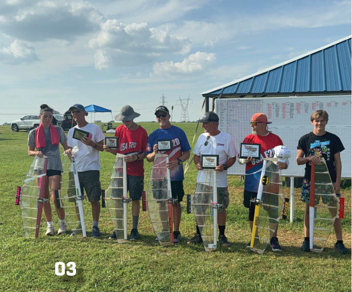 Seven people holding model planes and plaques on a grassy field.
