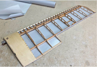Model airplane wing with wooden frame and translucent covering, on a workshop floor.