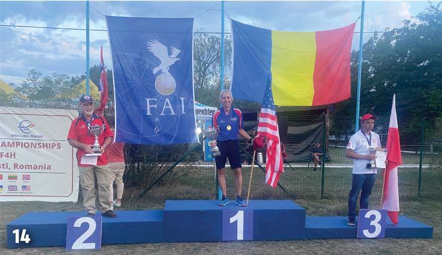 Three men holding trophies stand on podiums numbered 1, 2, and 3 under flags.