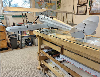 Model biplane on a wooden desk in a room with maps and framed pictures.