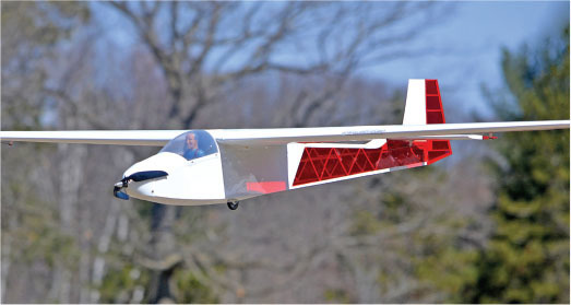White and red model glider flying outdoors with trees in the background.