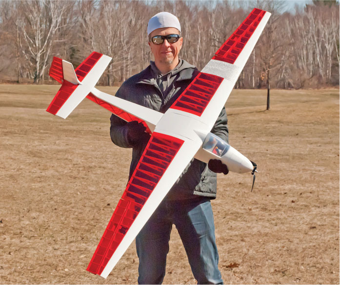 Man holding a red and white model airplane in a grassy field.