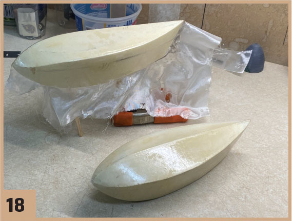 Two fiberglass model boat hulls on a workbench, background cluttered. Number 18 in corner.