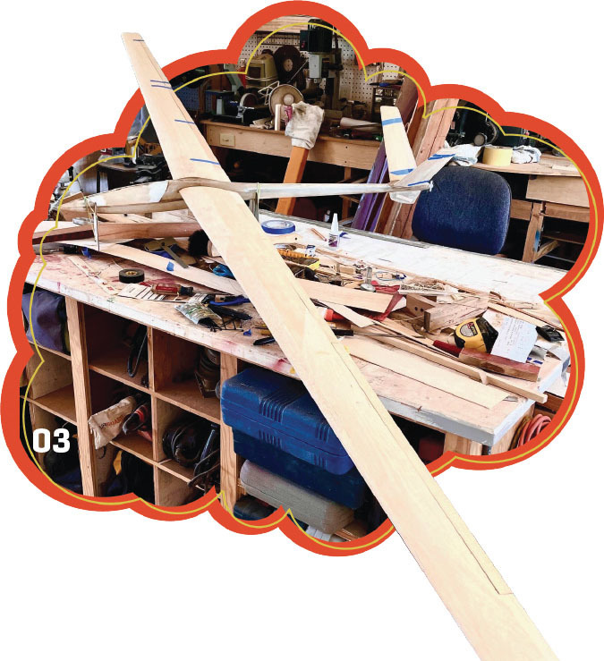 Wooden glider model on cluttered workshop table with tools and shelves.