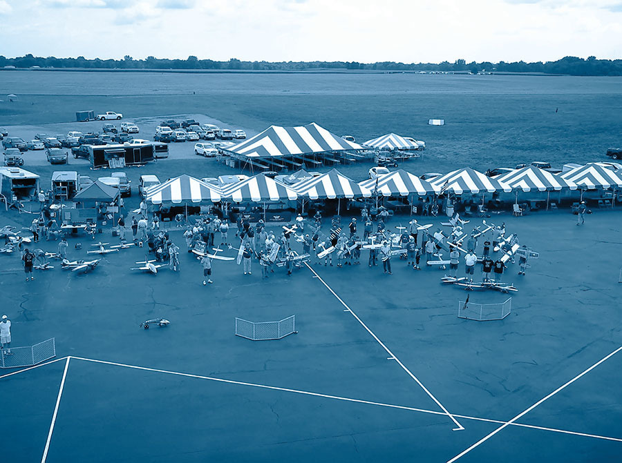 Outdoor event with model planes, striped tents, and crowd on an airstrip.