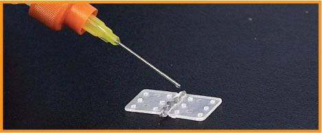 A needle approaches a small, transparent hinge on a dark surface.