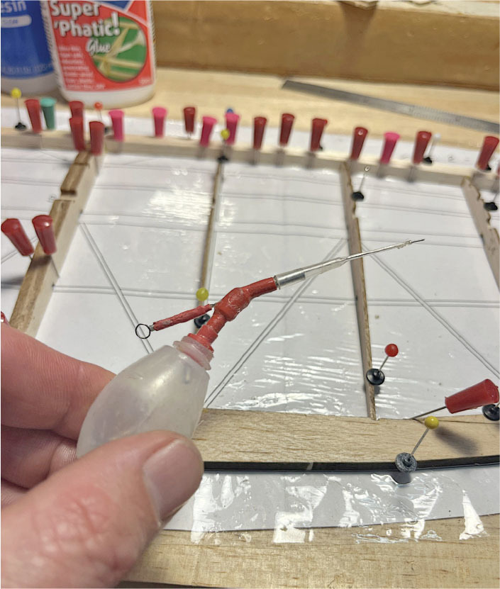 Hand holding glue bottle near model airplane frame with pins and lines.