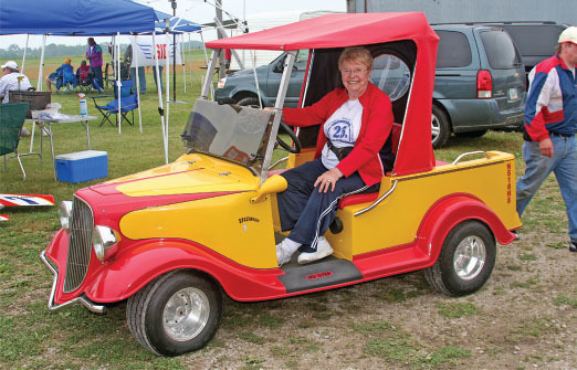 Smiling woman in a red and yellow retro-style golf cart at an outdoor event.