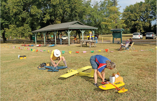 Two people working on model airplanes in a grassy park near a pavilion.