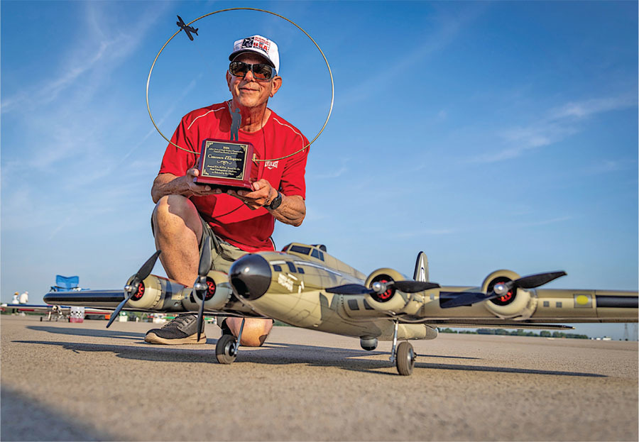 Man holding plaque, crouching by model airplane on runway, blue sky background.