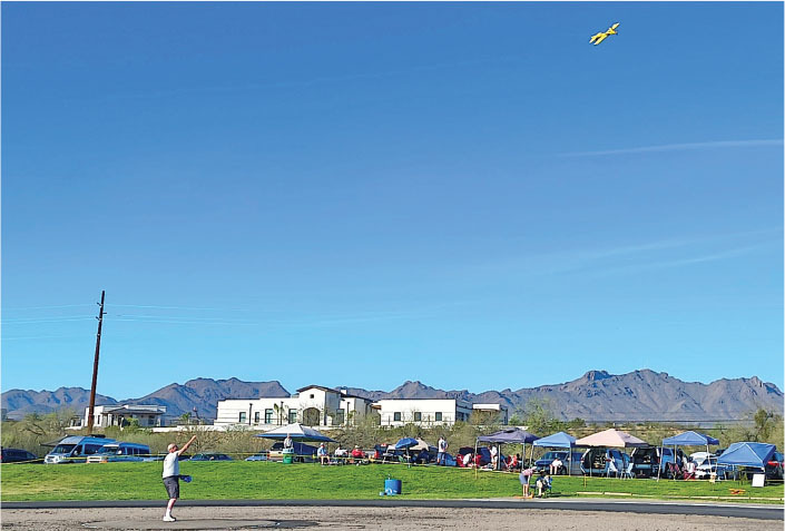 Person flying a yellow model airplane in a park with tents and mountains.