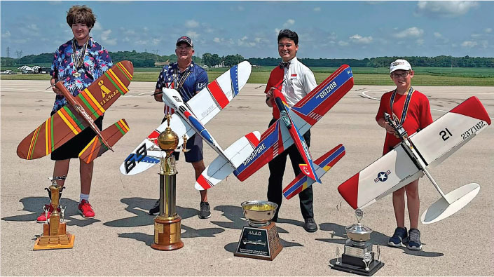 Four people holding model planes with trophies on a runway.