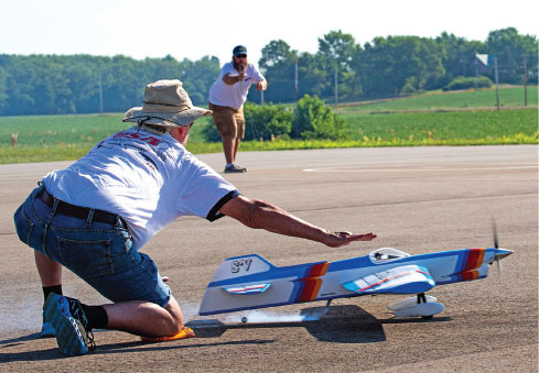 Two men launching a small model aircraft on a sunny day, with fields in the background.