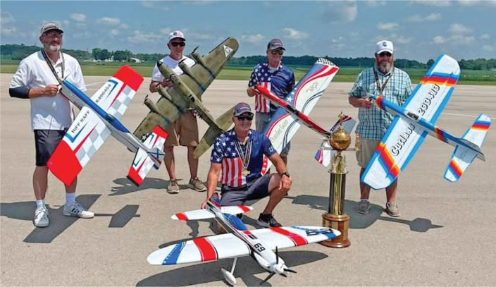 Five men pose with model planes and a trophy on an airfield.