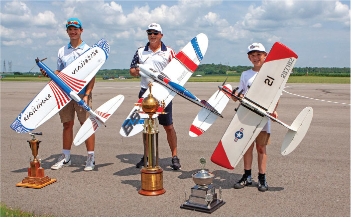 Three people with model planes and trophies on an airstrip.