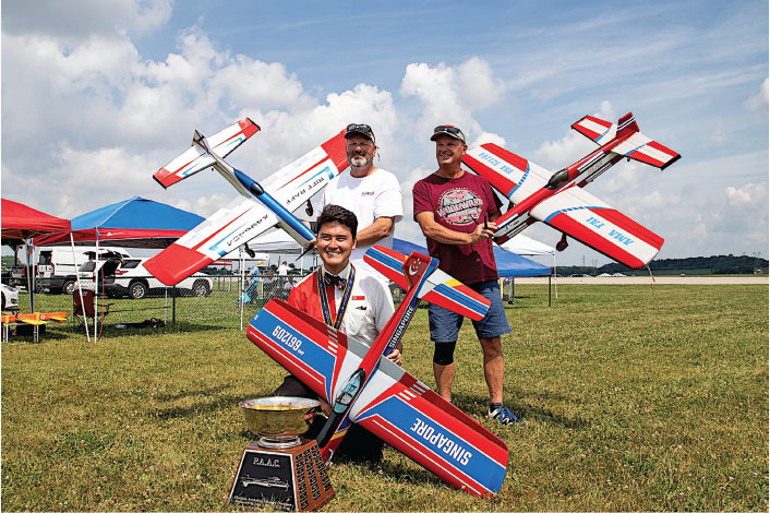 Three men holding model airplanes at an outdoor event, one kneeling with a trophy.