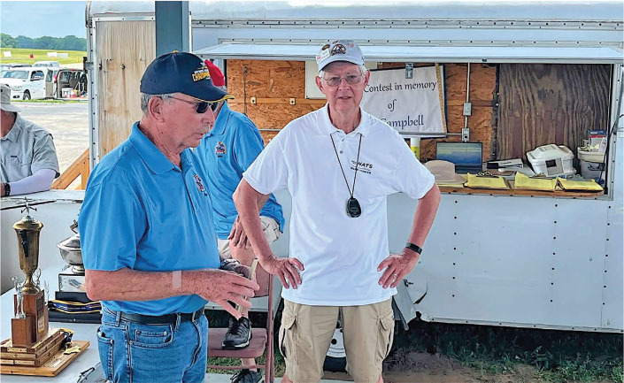 Two men in hats at an outdoor event, standing near a trailer.