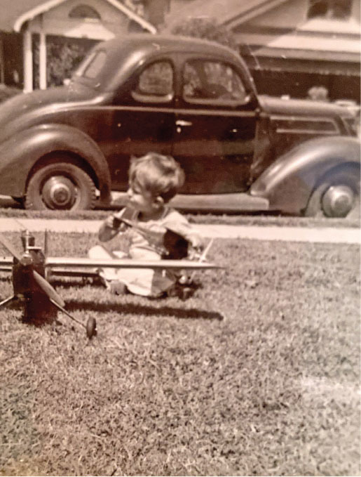 Child playing with a toy plane on grass, vintage car in background.