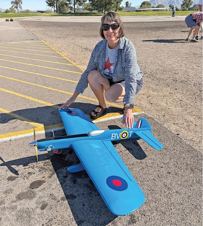 A person squats beside a large blue model airplane on asphalt.