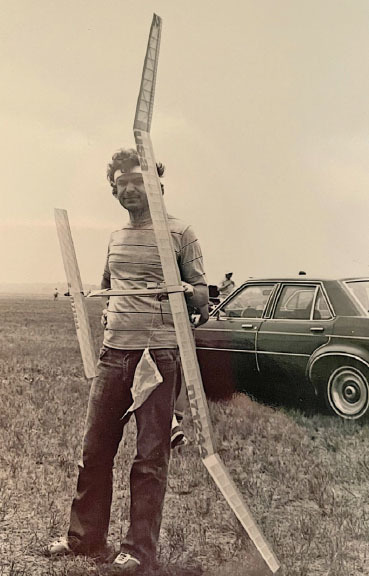 Man holding a model airplane stands beside a car in a grassy field, black and white photo.