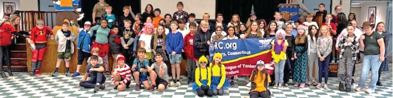 A large group of children in costumes stand on a checkered floor in front of a blue banner.
