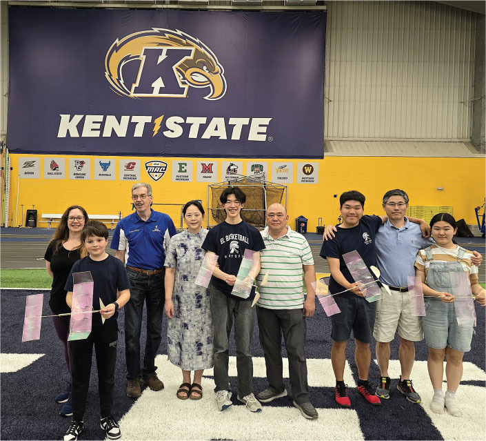 Nine people holding kites, standing in front of a Kent State banner indoors.