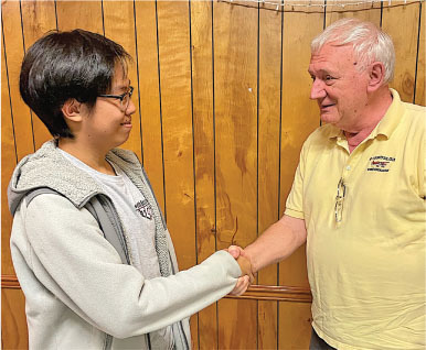 Two people shaking hands in front of a wooden wall, smiling at each other.
