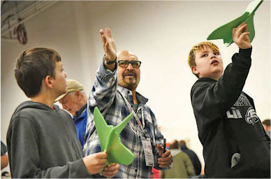 Boys and a man hold green foam planes indoors, smiling and enthusiastic.