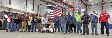 Group of people holding model airplanes in a large hangar with an American flag.